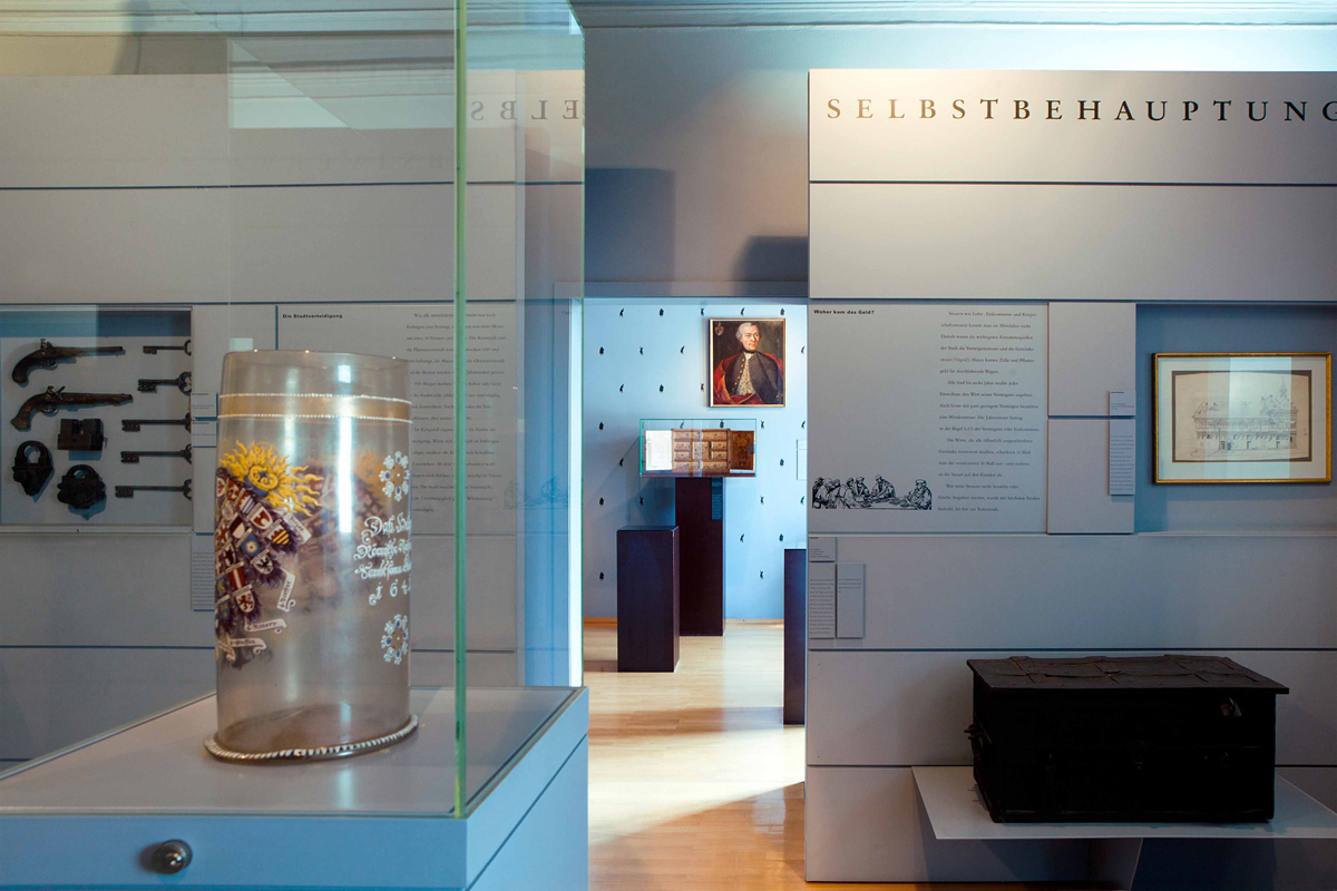 Glaskrug Zwei Räume, die von einem offenen Durchgang getrennt sind. Im vorderen Raum steht in einer Vitrine ein großer bemalter Glaskrug, der sogenannte Reichsadler-Humpen, weiter hinten eine große Truhe, die Geldkasse.