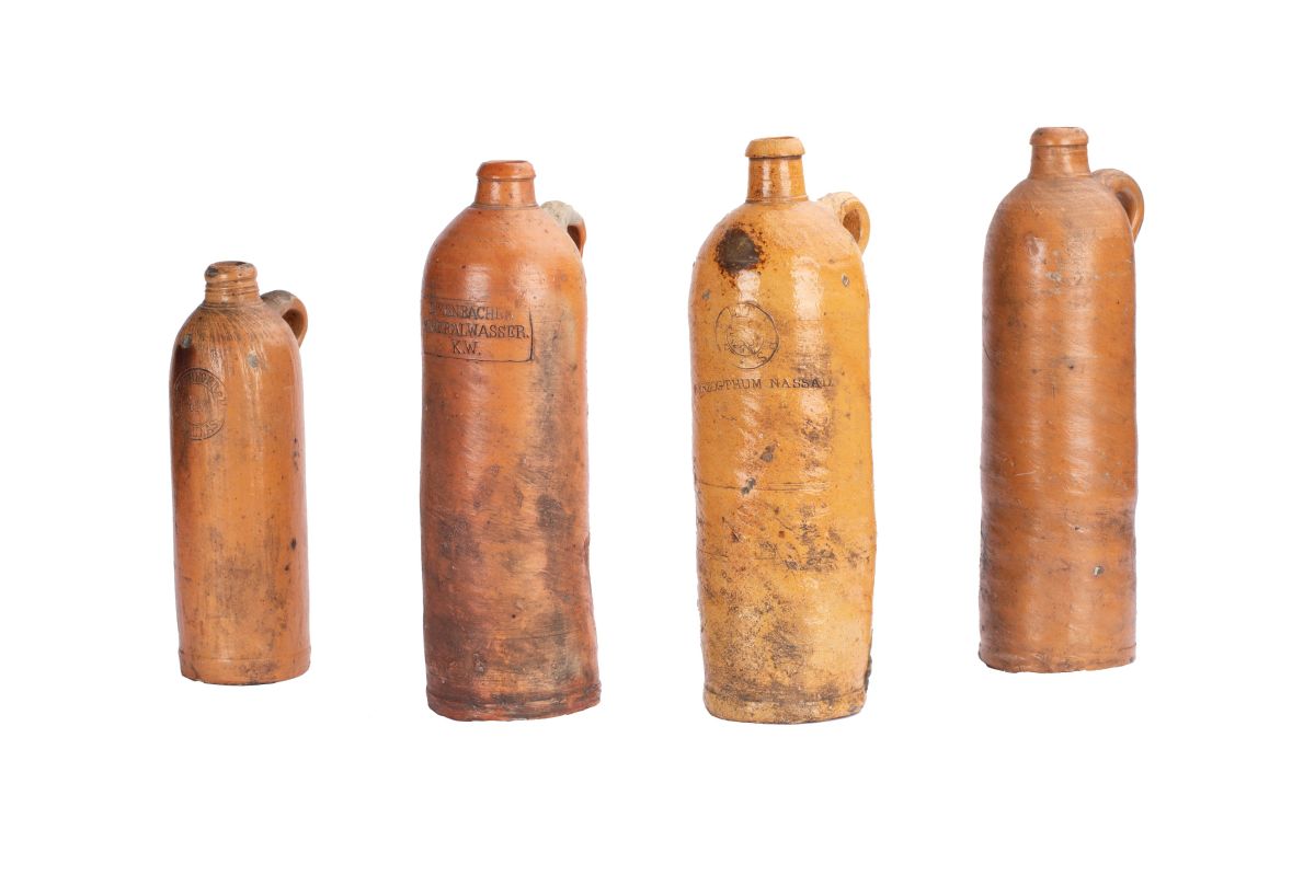 Vier hellbraune Flaschen mit schmalen Hals und Henkel. Auf jeder Flasche ist ein Stempel des jeweiligen Quellortes aufgedruckt. Vier hellbraune Flaschen mit schmalen Hals und Henkel. Auf jeder Flasche ist ein Stempel des jeweiligen Quellortes aufgedruckt.