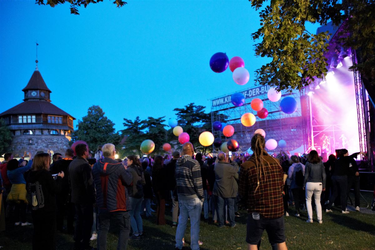 Konzert auf der Esslinger Burg © Kulturamt/ Fretwurst Viele Menschen stehen im Grünen vor einer großen Open-Air-Bühne, der Himmel ist abendblau. Über den Menschen sind bunte Luftballons zu sehen. Im Hintergrund erkennt man den Dicken Turm der Esslinger Burg