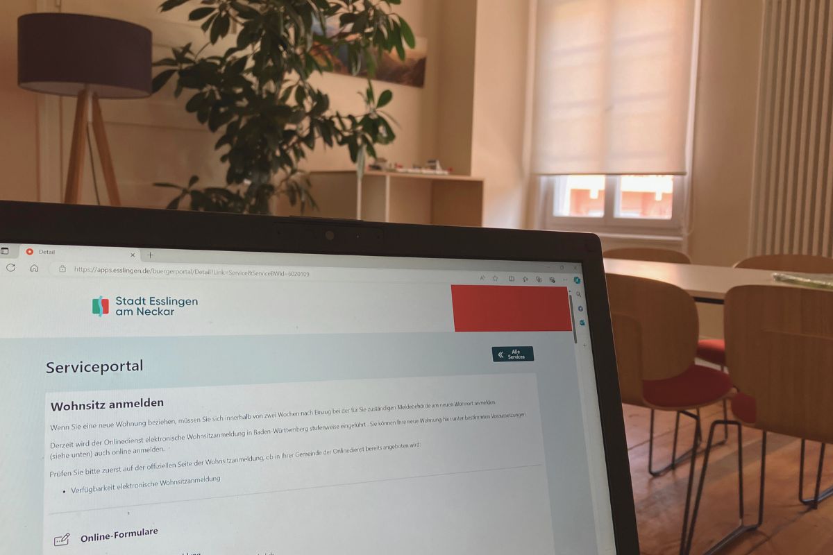 Digitale Wohnsitzanmeldung Das Online-Serviceportal auf einem Laptop, im Hintergrund ein Wohnzimmer