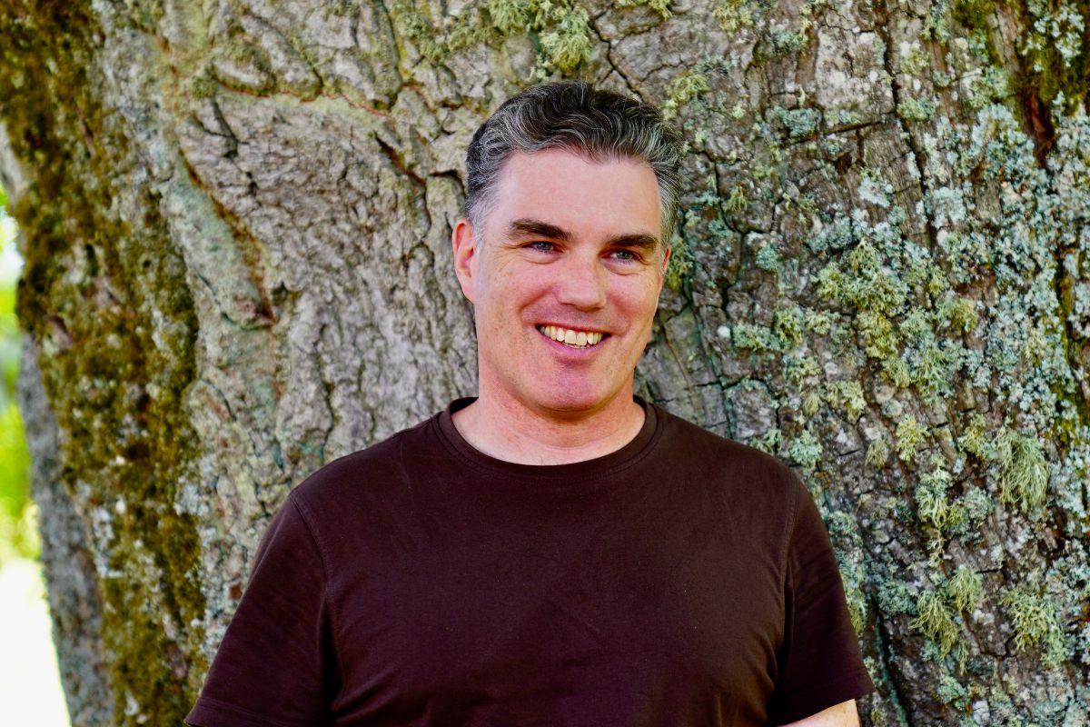 Christian Schwenk steht im braunen T-Shirt vor einem Baum und lächelt in die Kamera.