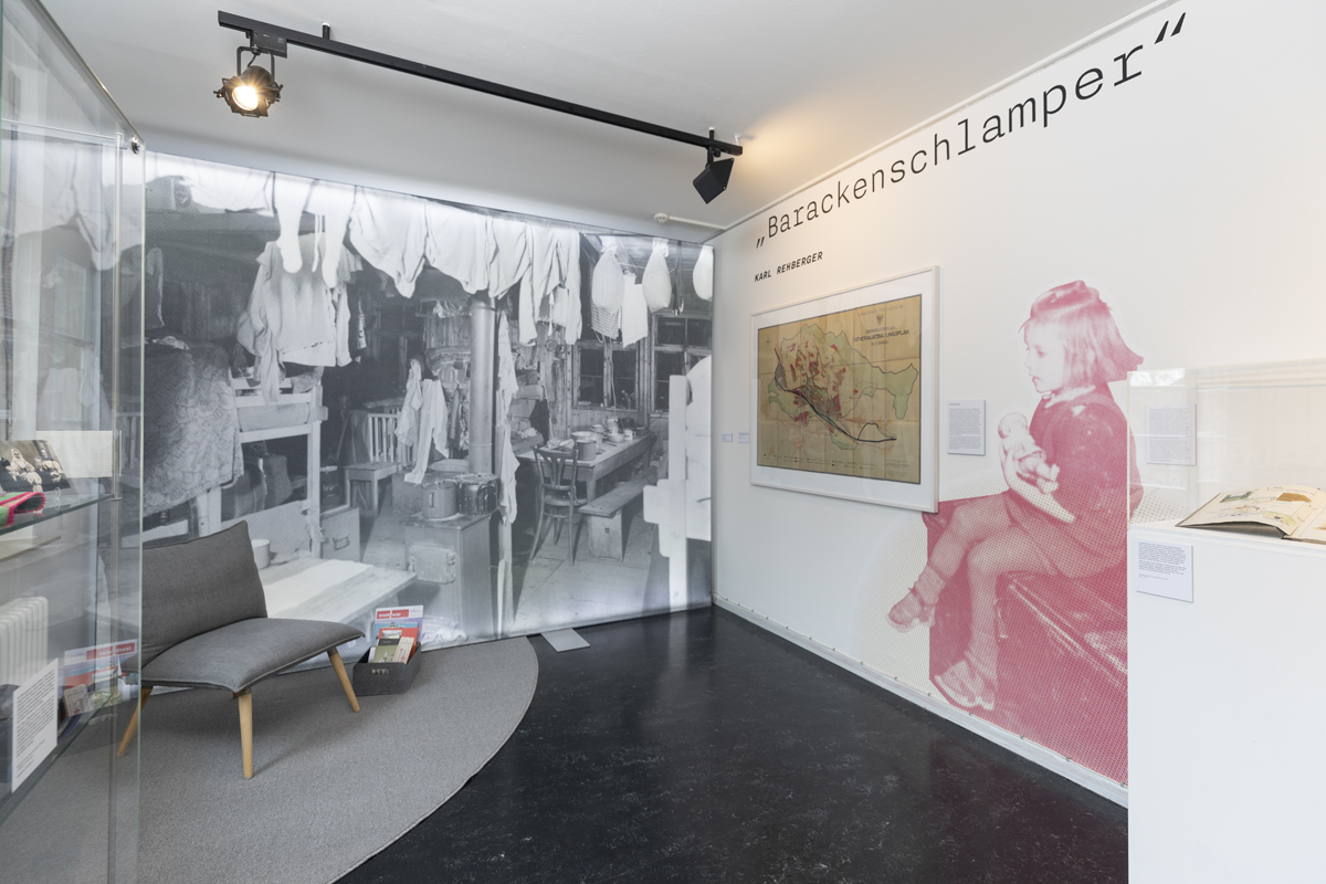 An der Wand ein riesiges Foto eines kleinen Mädchens, das auf einer Kiste sitzt und eine Puppe im Arm. An der Rückwand ein wandfüllendes Foto aus einer baracke.