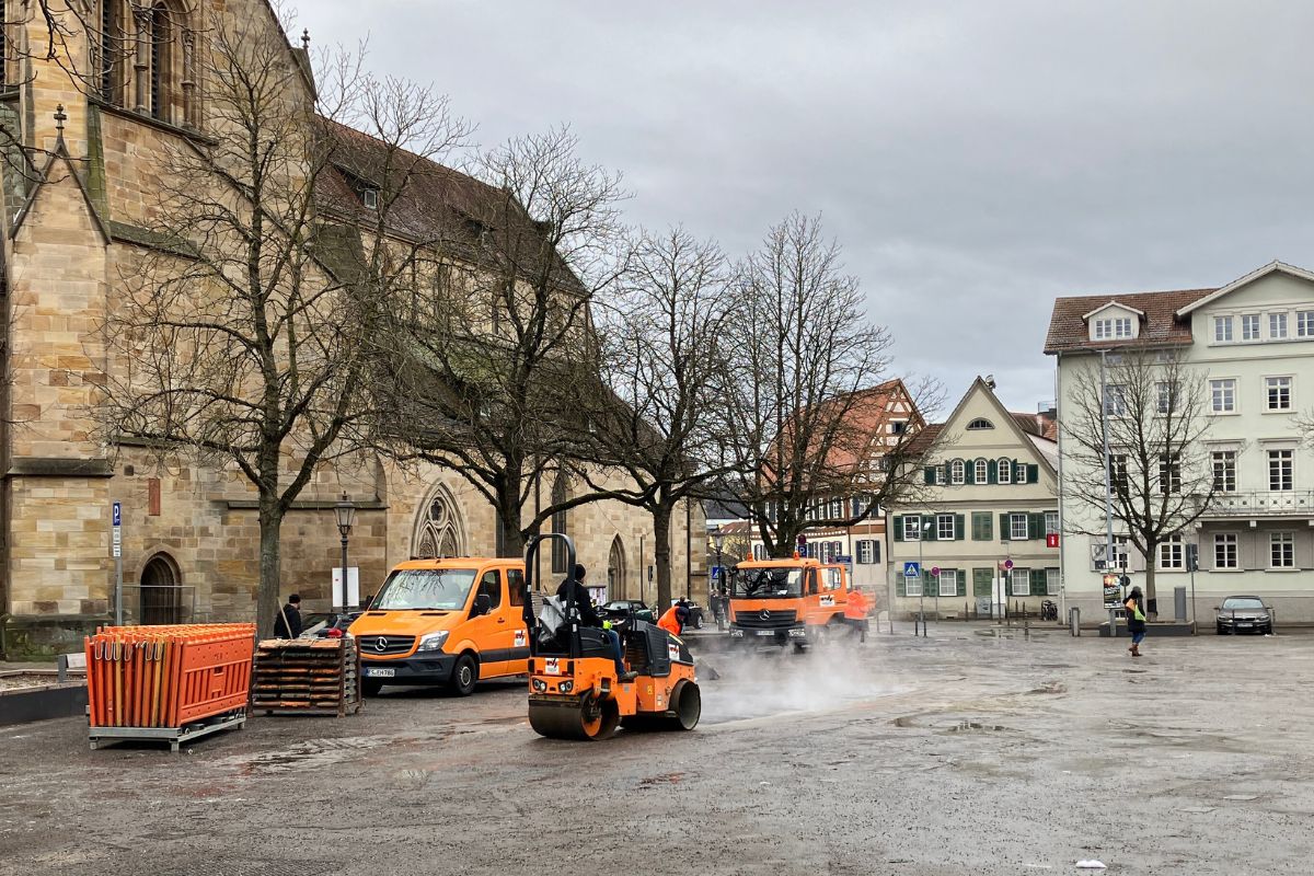 Baufahrzeuge asphaltieren die Stellen, an denen der Marktplatz aufgegraben wurde. Im Hintergrund sind die Stadtkirche und die Abt-Fulrad-Straße zu sehen.