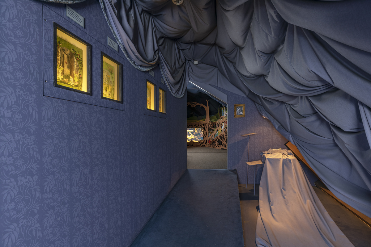 Im Schreiber-Museum: Schmaler dunkler Gang, links sind kleine Schaukästen an der Wand, rechts liegt ein Rüschenkleid mit überlangem Rock.