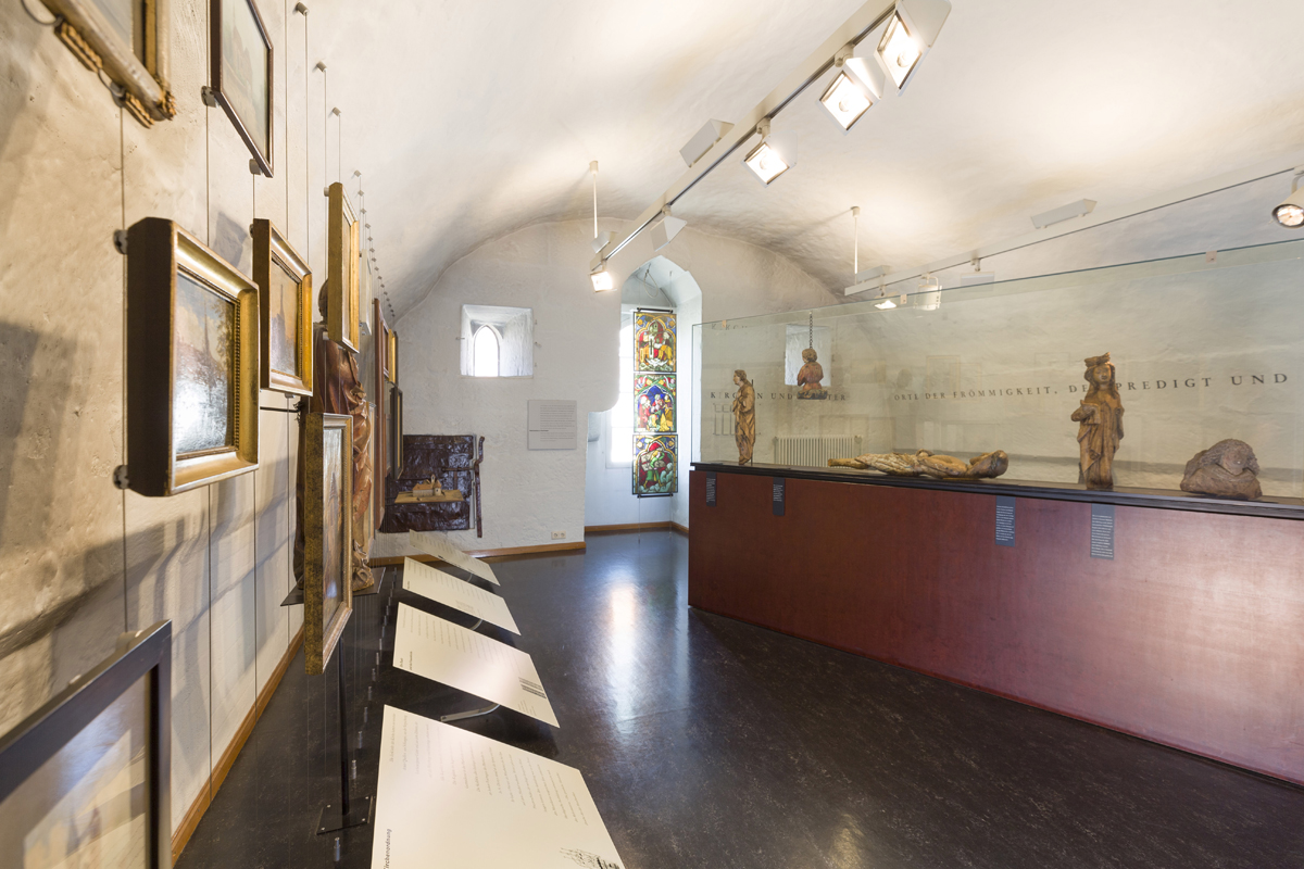 Ausstellungsraum: Ein Buntglasfenster und Gemälde an der Wand.