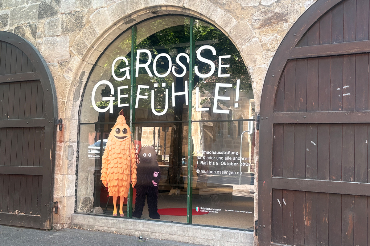 Fenster des Ausstellungsraumes. Großes Fenster, das mit dem Ausstellungstitel Große Gefühle und zwei Figuren beklebt ist.