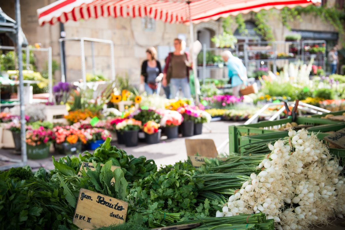 Marktstand mit Kräutern und Frühlingszwiebeln, im Hintergrund Blumen und Marktbesucher.