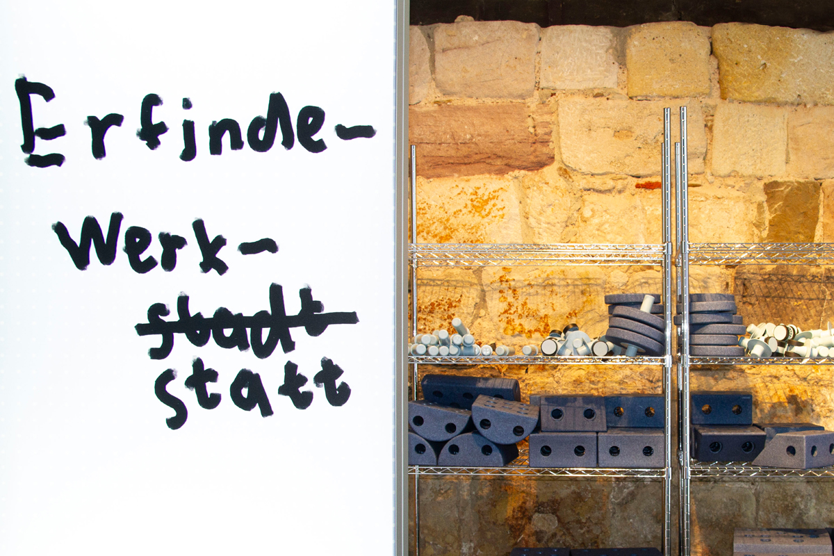 Hinter einem Schild "Erfindewerkstatt" steht ein Regal mit verschiedenen Bauteilen.