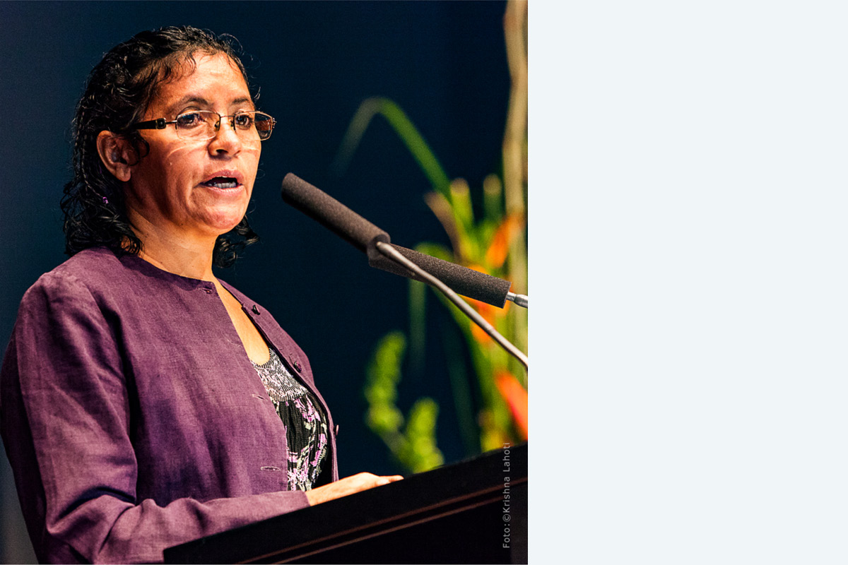 Frau mit dunklen Locken und Brille in violetter Jacke spricht am Rednerpult in ein Mikrofon