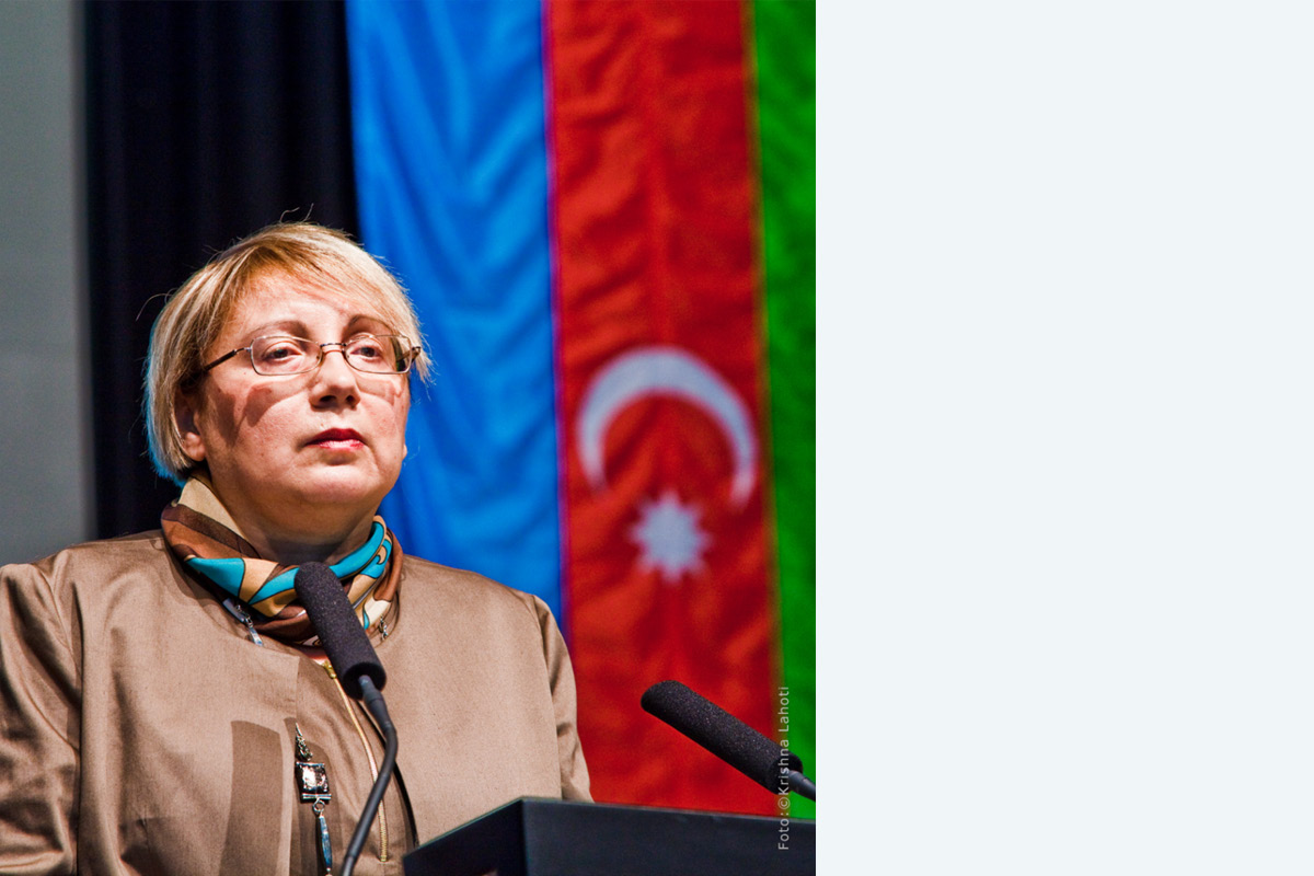 Frau mit kurzen, blonden Haaren und Brille in brauner Jacke mit Halstuch spricht am Rednerpult in ein Mikrofon. Im  Hintergrund eine Flagge mit blauem, rotem und grünem Streifen. Auf dem roten sind ein Halbmond und ein Stern zu sehen.