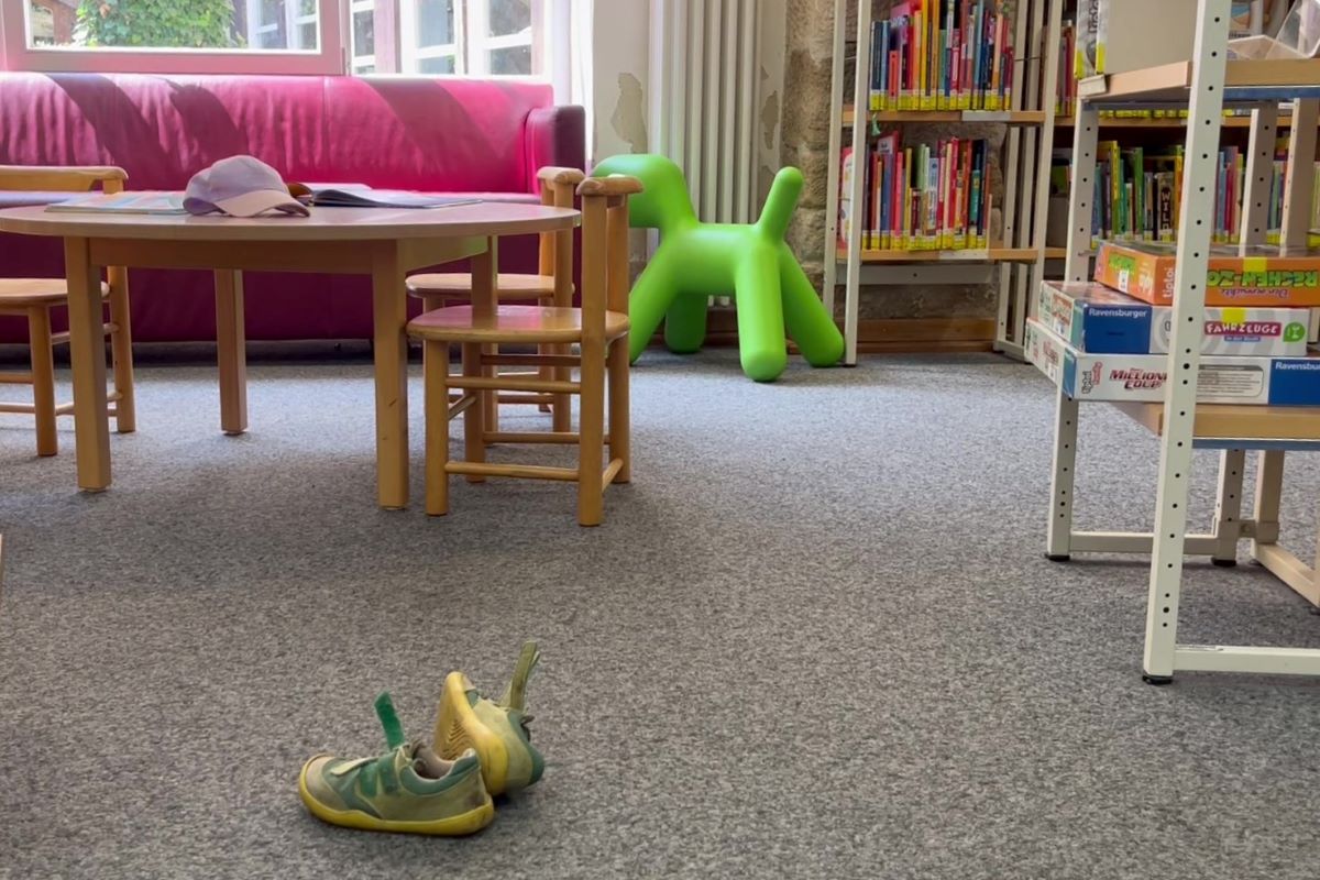 Kinderbereich mit Tisch, kleinen Stühlen, roten Sofa, grüner Spielfigur und einem Paar Kinderschuhe auf dem Teppich