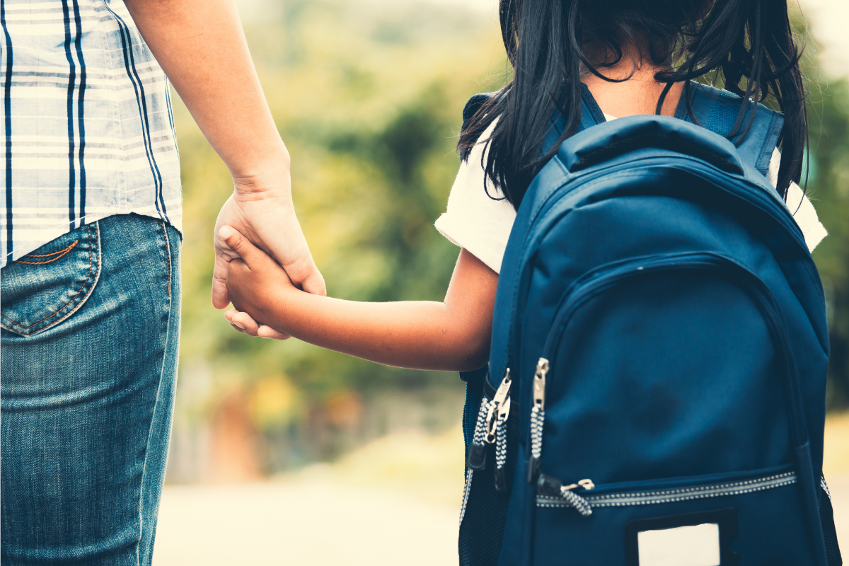 Links läuft ein Elternteil und hält ein Mädchen an der Hand, das rechts läuft. Beide sind auf einem Gehweg auf dem Weg zur Schule.