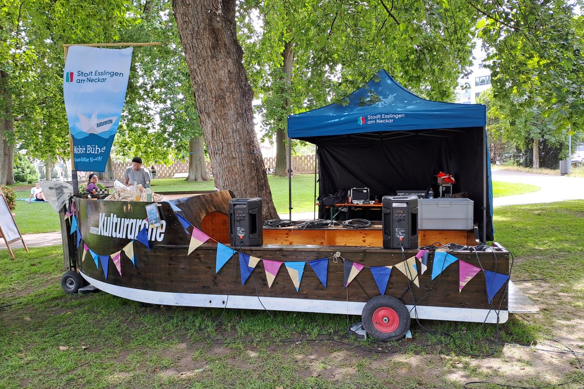Kulturarche beim Schwörfest 2024, Foto: © Kulturamt Esslingen Ein mit Wimpeln und Fahne geschmücktes Holzbook steht im Grünen. Auf dem Boot steht "Kulturarche". Im Hintergrund steht ein blauer Pavillon.