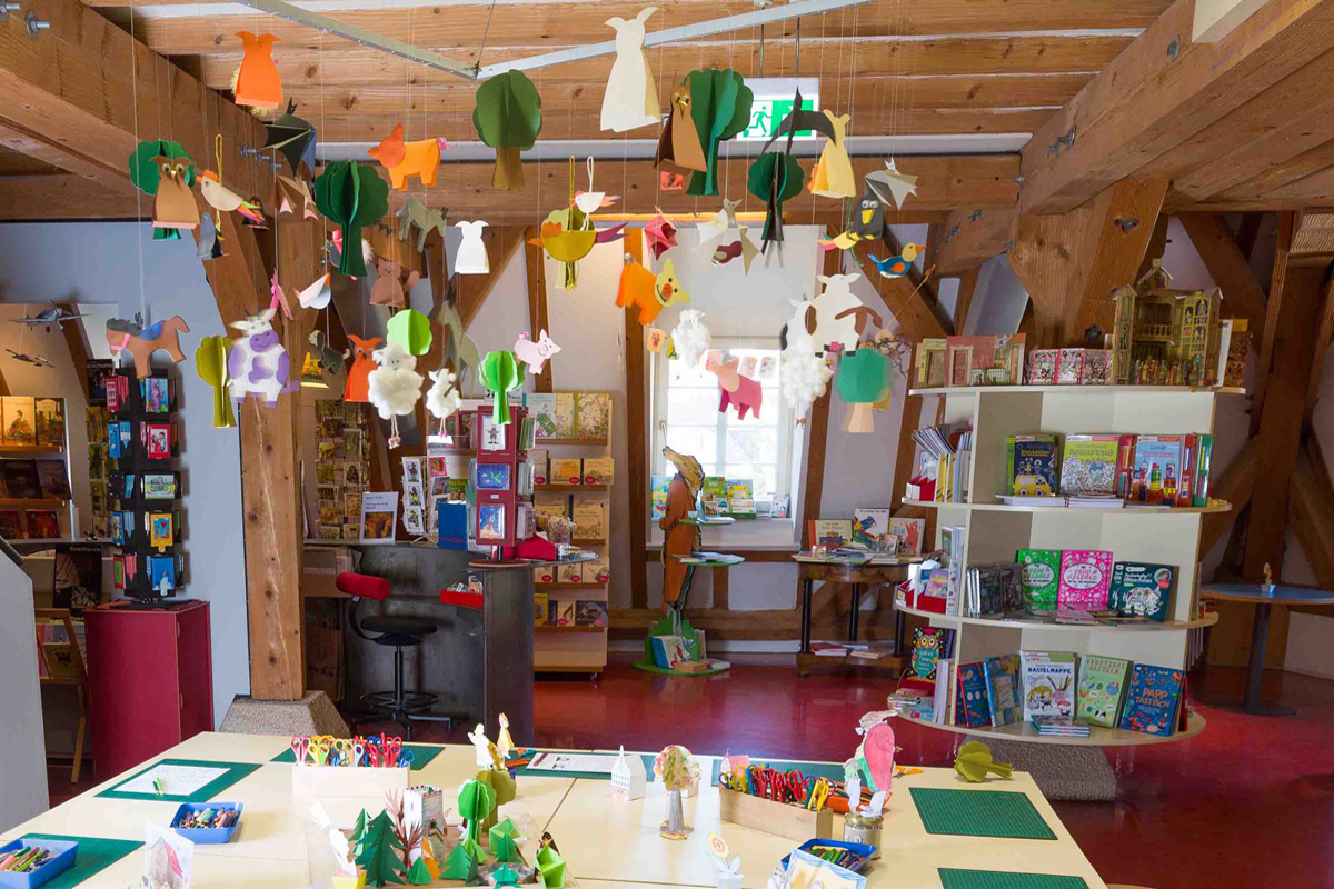 Fotografie: Michael Saile (Ausschnitt). Basteltisch mit Bastelbeispielen, im Hintergrund der Shop-Bereich mit Bücherregalen.