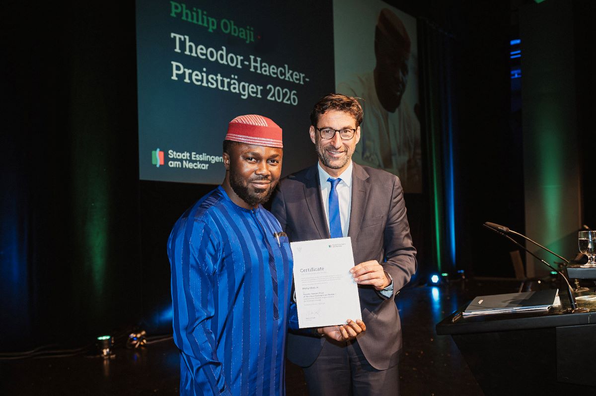 Zwei Männer stehen auf einer Bühne und halten gemeinsam eine Urkunde. Im Hintergrund ist eine große Leinwand mit Text zur Preisverleihung „Theodor-Haecker-Preisträger 2026“ zu sehen.