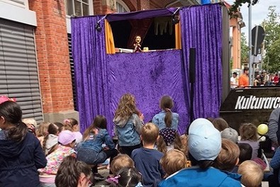 Auf dem schwarzen Holzboot "Kulturarche" ist eine Bühne mit lila Vorhang aufgebaut. Ein Kasper ist zu sehen und viele Kinder, die vor der Kulturarche sitzen und stehen.