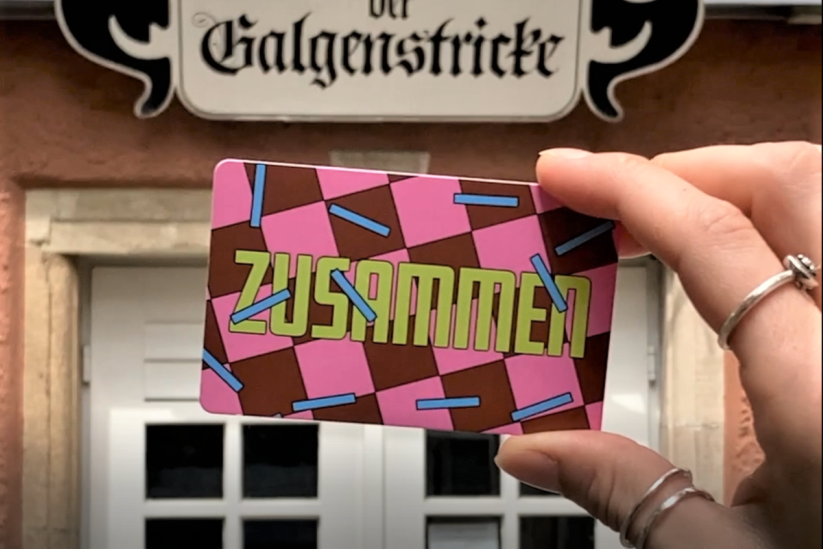 Eine Hand zeigt eine Kulturpass-Skonto-Karte im Scheckkartenformat. Die Karte ist rosa-braun-kariet, die Aufschrift in grün heißt "ZUSAMMEN". Im Hintergrund erkennt man den Eingang zum Kabarett der Galgenstricke in der Esslinger Webergasse.