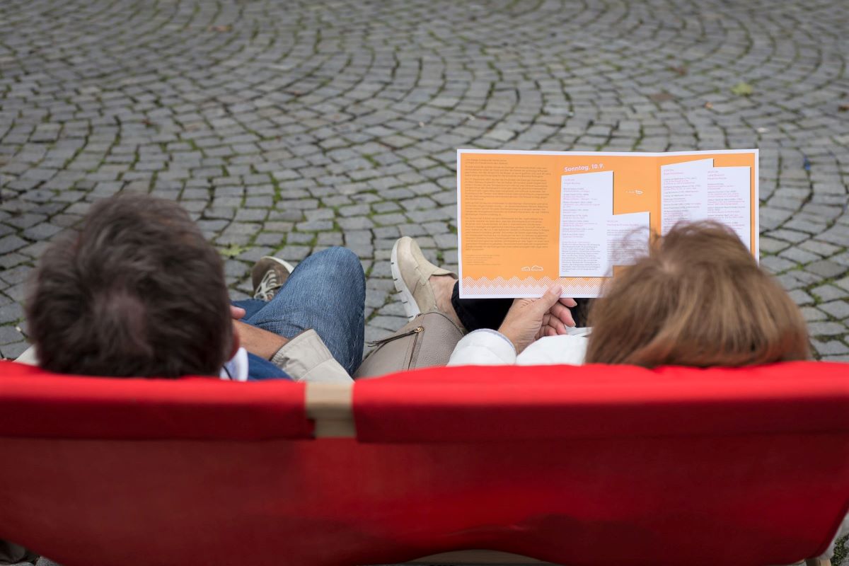 Ein Paar liest das Programmheft des Glockenspiel-Festivals in einem roten Liegestuhl