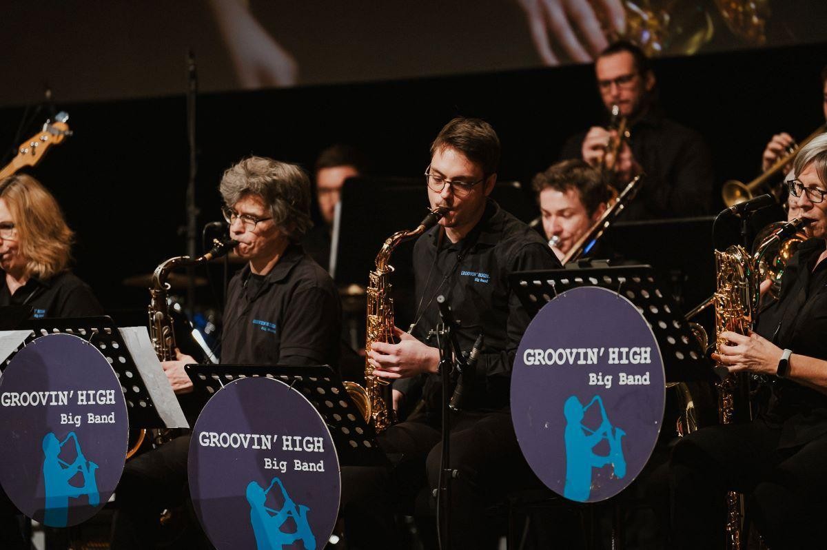 Groovin' High Big Band Mehrere Musiker:innen einer Big Band spielen Saxophon und Trompete auf einer Bühne. Sie tragen schwarze Hemden mit dem Bandnamen, vor ihnen stehen Notenständer mit blauen Logos