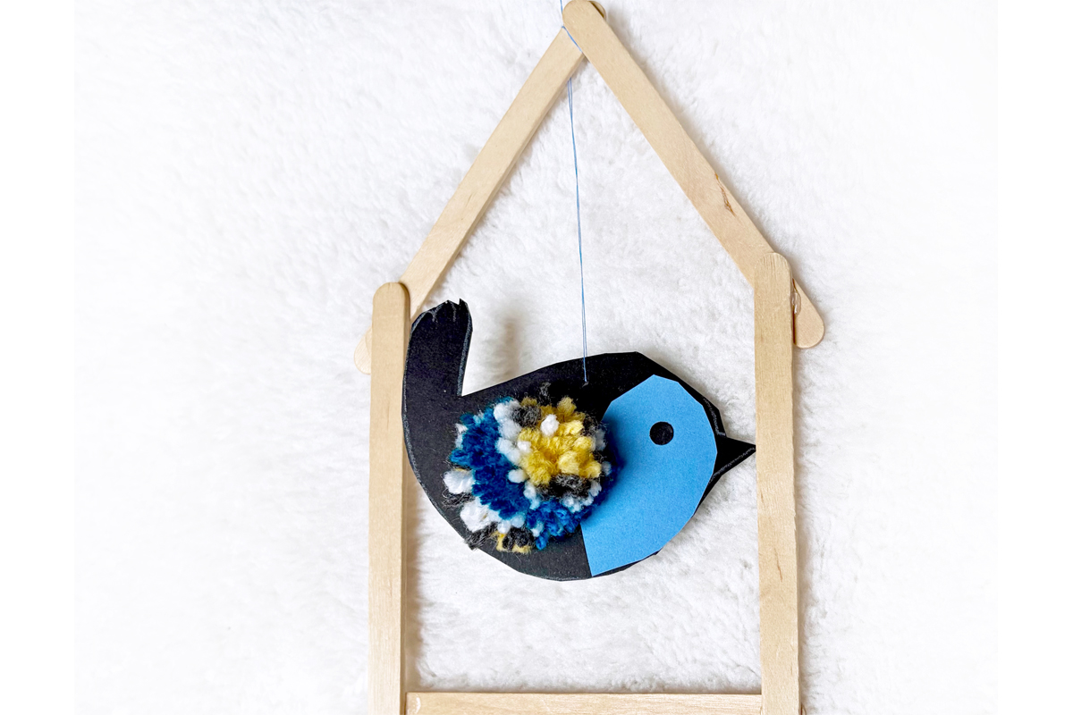EIn Vogel ist aus farbigem Papier gebastelt und verziert. Er hängt in einem Rahmen aus Holzstäbchen.