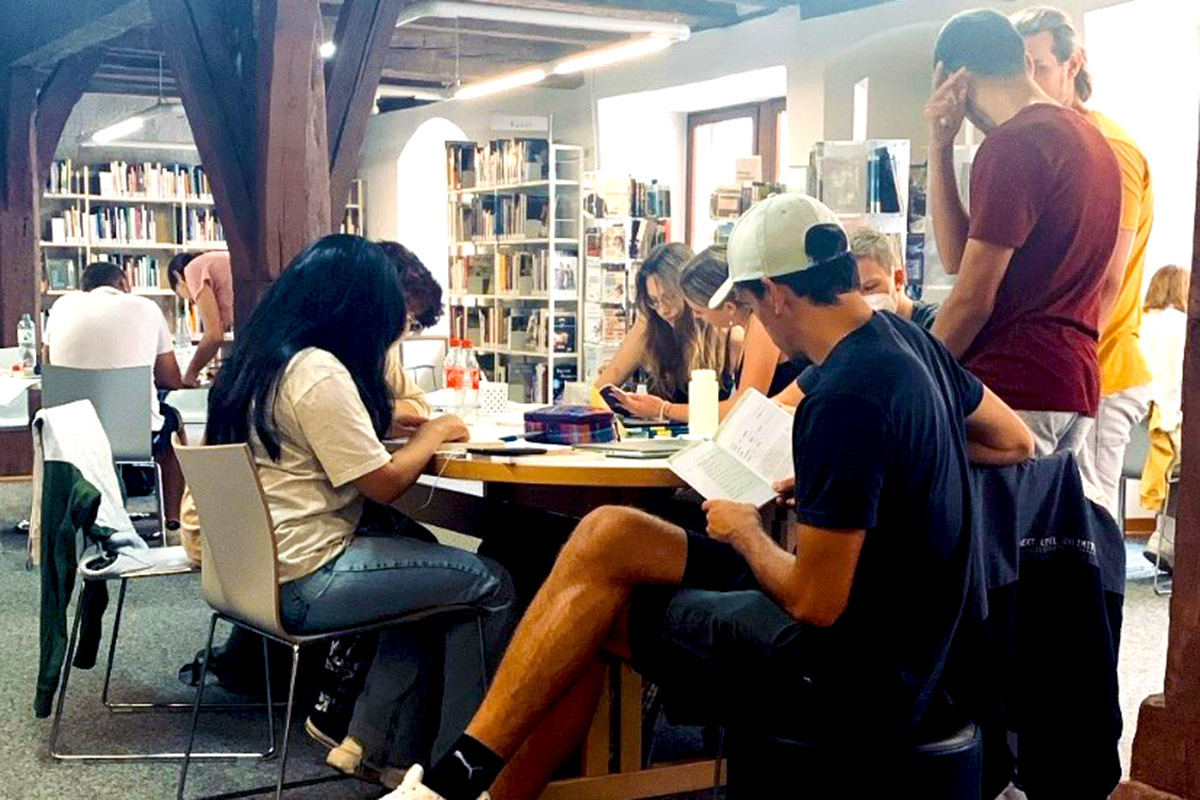 Junge Leute sitzen und stehen mit Lernmaterial ausgestattet um einen Tisch in einem Raum, an dessen Wänden Bücherregale stehen