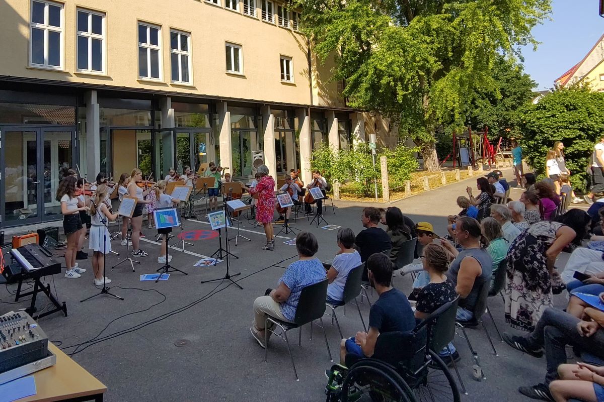 Serenade im Hof © Städtische Musikschule Esslingen Vor einem Gebäude im Schatten spielt ein Kinder-Streichorchester, gegenüber sitzt Publikum auf Stühlen.
