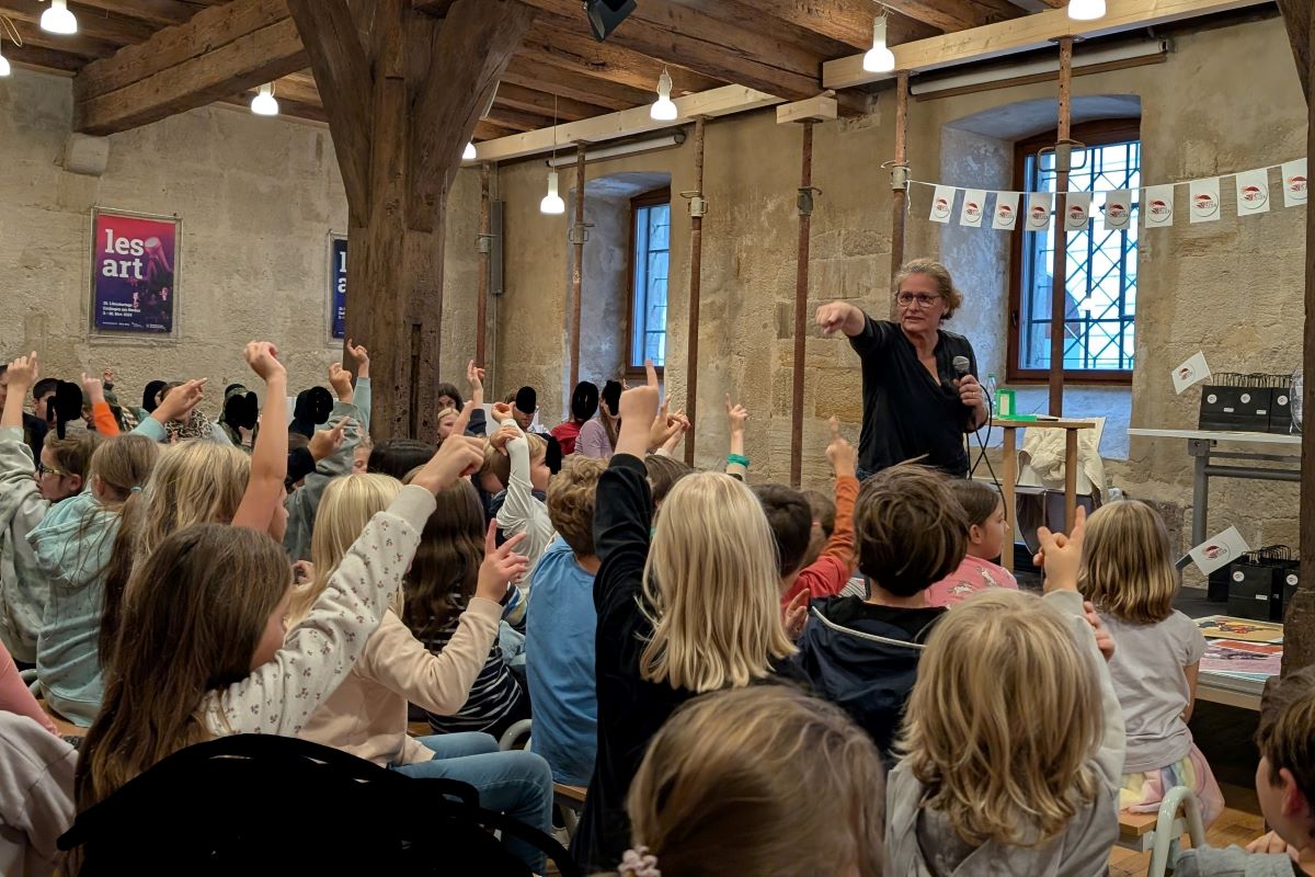 Viele sich meldende Kinder in einem Raum mit Holzbalken und Steinwänden