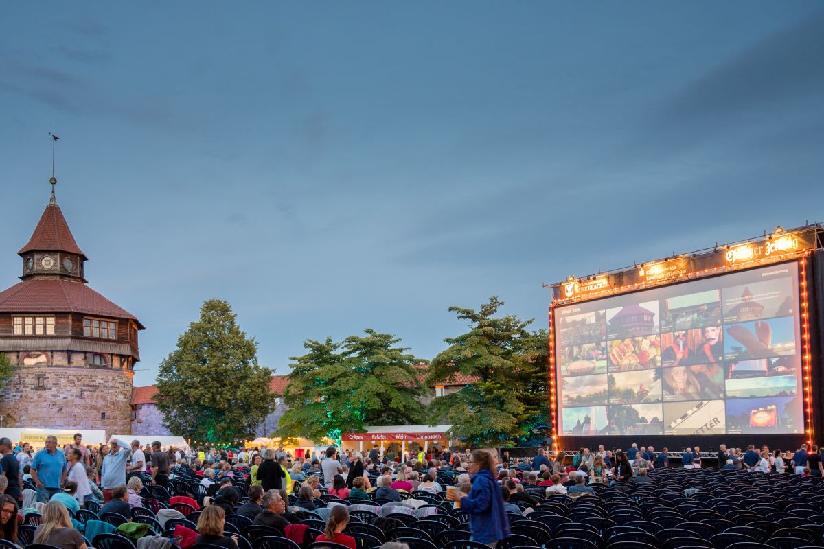 Eine große Filmleinwand steht im Freien, davor sitzen Menschen auf Stühlen und Picknickdecken. Links ist der Dicke Turm der Esslinger Burg zu sehen